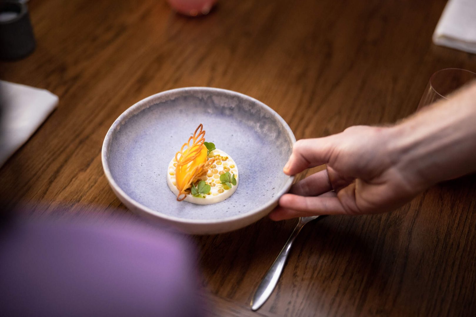 A hand holding a blue ceramic plate with a small amount of food artfully arranged and presented.